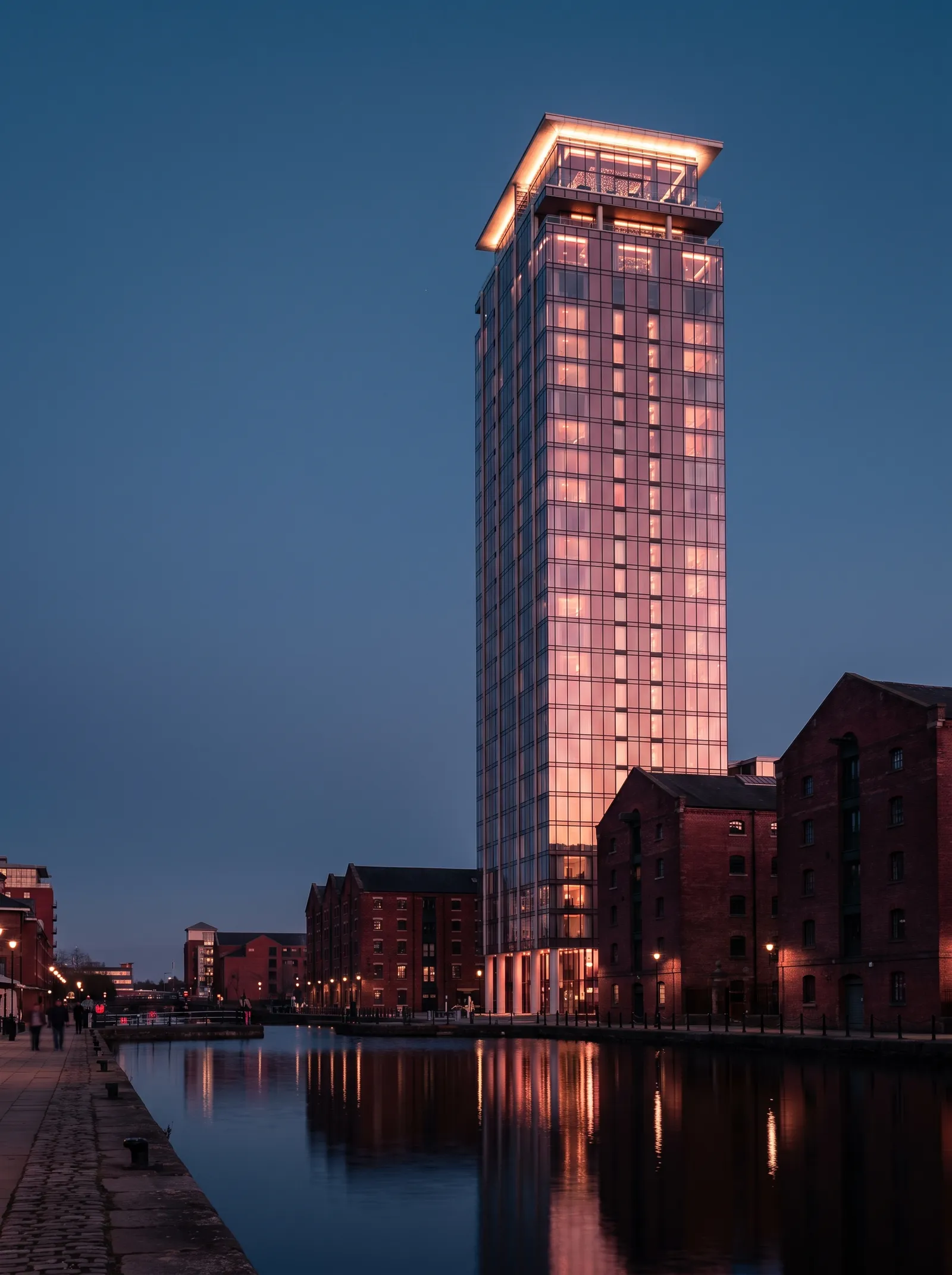 The Lumenquay glass tower glowing rose at blue hour above the canal basin and its brick warehouses